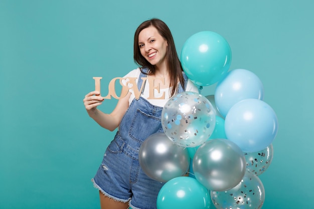 Lächelnde junge Frau in Denim-Kleidung, die Holzwortbuchstaben Liebe hält und mit bunten Luftballons feiert, die auf blauem türkisfarbenem Hintergrund isoliert sind. Geburtstagsfeier, Menschen Emotionen Konzept.