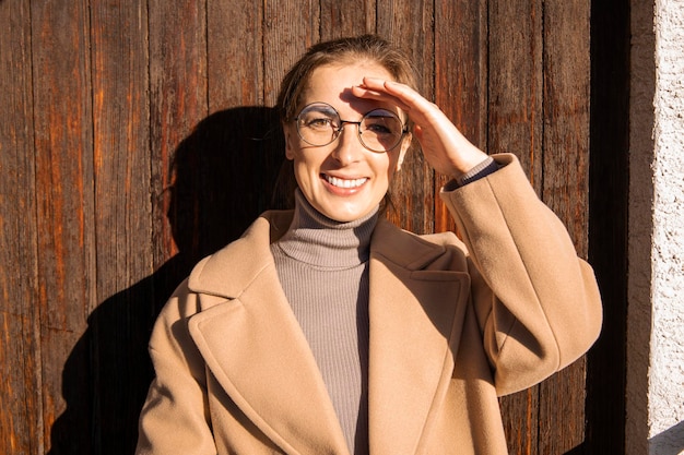 Lächelnde junge Frau in einem Mantel mit Brille an einem sonnigen Tag auf einem hölzernen Hintergrund