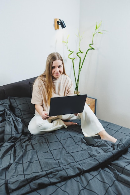 Lächelnde junge Frau in Freizeitkleidung, die lächelt und aus der Ferne arbeitet, sitzt auf dem Bett mit Laptop während der Fernarbeit Freiberufliche Arbeit Arbeiten Sie von zu Hause aus