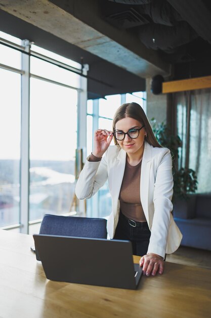 Lächelnde junge Frau in Freizeitkleidung und Brille, die eine weiße Jacke trägt und lacht, während sie mit einem Laptop an einem Tisch sitzt, während sie arbeitet Arbeiten im Büro