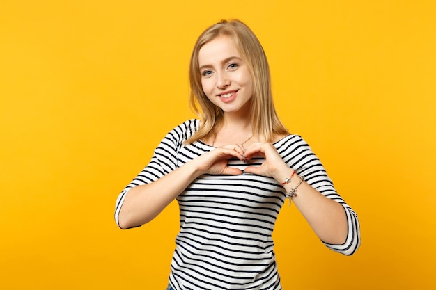 Lächelnde junge Frau in gestreifter Kleidung, die eine Kamera zeigt, die Formherz mit Händen zeigt, die auf gelb-orangefarbenem Wandhintergrund im Studio isoliert sind. Menschen aufrichtige Emotionen Lifestyle-Konzept. Mock-up-Kopienbereich.