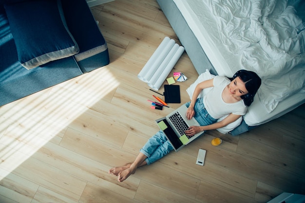 Lächelnde junge Frau in Jeans und T-Shirt, die mit einem Laptop und Schreibwaren auf dem Boden sitzt Website-Banner