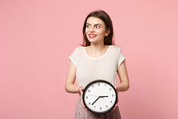 Lächelnde junge Frau in lässiger leichter Kleidung posiert isoliert auf pastellrosa Wandhintergrund, Studioportrait. People-Lifestyle-Konzept. Mock-up-Kopienbereich. Hält die Uhr in den Händen und schaut zur Seite.