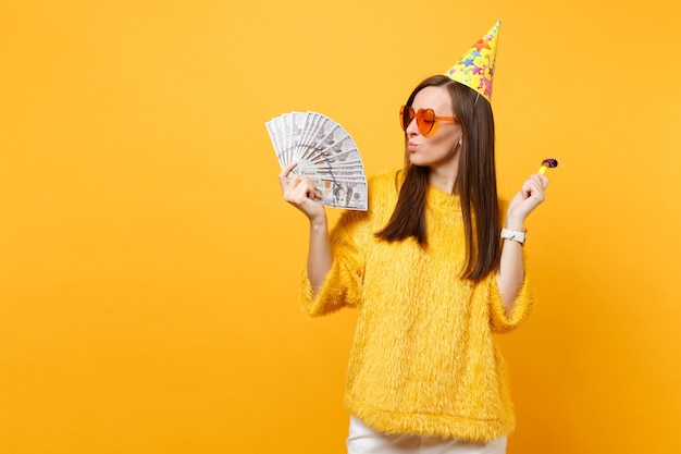 Lächelnde junge Frau in orangefarbener Herzbrille, Geburtstagshut mit Pfeife, die auf Bündel viele Dollar schaut, Bargeld, das auf gelbem Hintergrund einzeln feiert. Menschen aufrichtige Emotionen, Lebensstil.
