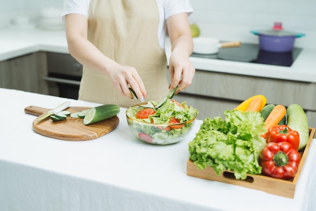 Lächelnde junge Frau in Schürze steht in moderner Küche und bereitet Salat zu