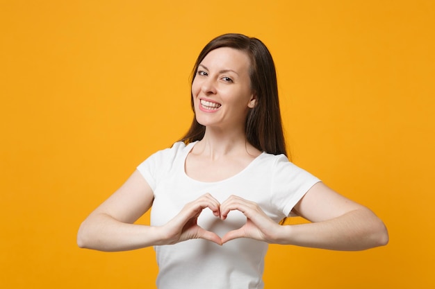 Lächelnde junge Frau in weißer Freizeitkleidung, die in die Kamera blickt und Formherz mit den Händen zeigt, herzförmiges Schild isoliert auf gelb-orangefarbenem Wandhintergrund. People-Lifestyle-Konzept. Mock-up-Kopienbereich.