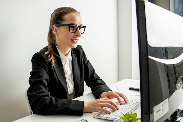 Lächelnde junge Frau mit Brille arbeitet, während sie am Arbeitsplatz an einem Computer sitzt