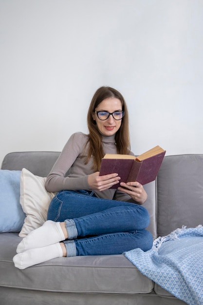 Lächelnde junge Frau mit Brille, die auf dem Sofa sitzt und ein Buch liest