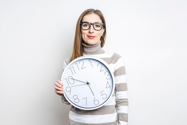 Lächelnde junge Frau mit Brille, die eine weiße Wanduhr auf hellem Hintergrund hält