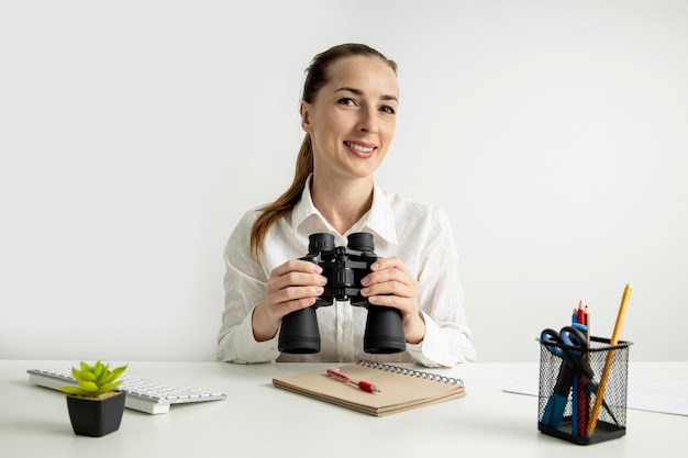 Lächelnde junge Frau mit dem Fernglas, die am Schreibtisch auf weißem Hintergrund sitzt