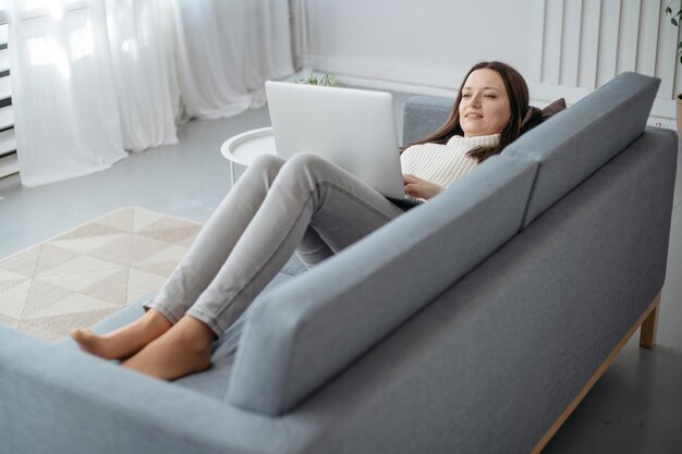 Lächelnde junge Frau mit einem Laptop, der auf dem Sofa im Wohnzimmer liegt