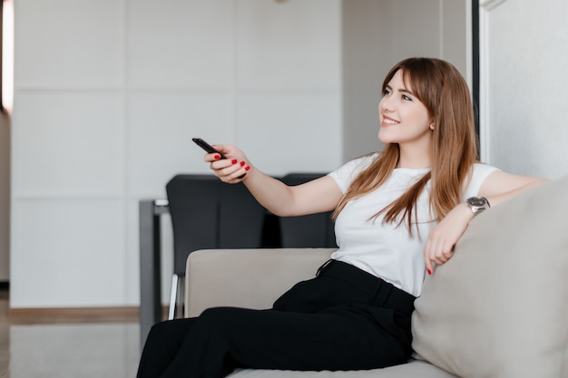 Lächelnde junge Frau mit Fernbedienung vom Fernsehen, das auf einer Couch zu Hause in der Wohnung sitzt
