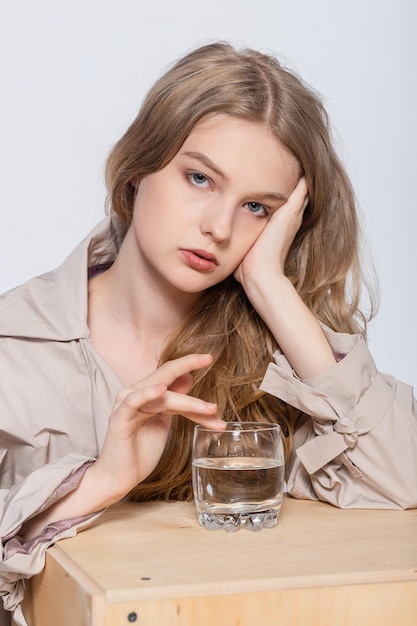 Lächelnde junge Frau mit Glas Wasser schönes Mädchen im beigen Regenmantel, der im Studio aufwirft