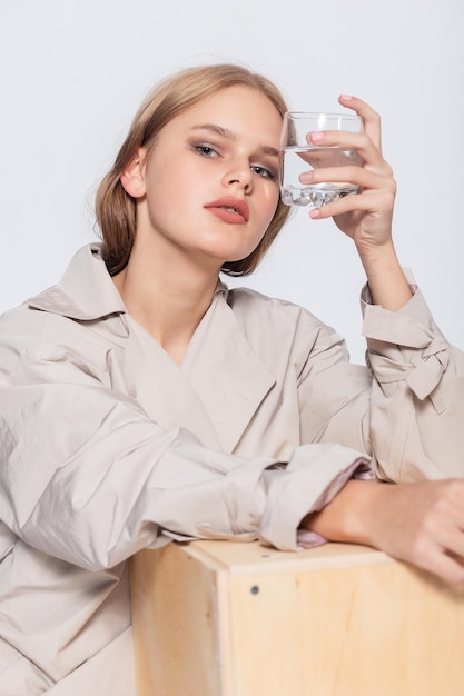 Lächelnde junge Frau mit Glas Wasser schönes Mädchen im beigen Regenmantel, der im Studio aufwirft