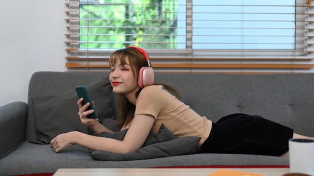 Lächelnde junge Frau mit Kopfhörer und Handy auf der Couch im hellen Wohnzimmer