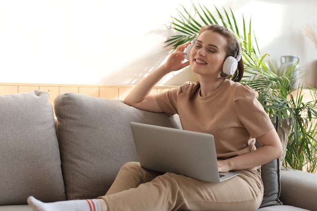 Lächelnde junge Frau mit Kopfhörern und Laptop auf dem Sofa.