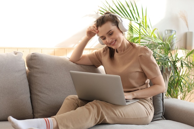 Lächelnde junge Frau mit Kopfhörern und Laptop auf dem Sofa.