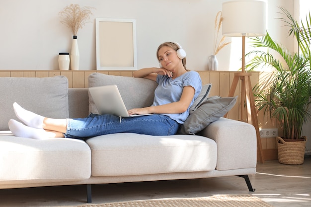 Lächelnde junge Frau mit Kopfhörern und Laptop auf dem Sofa.