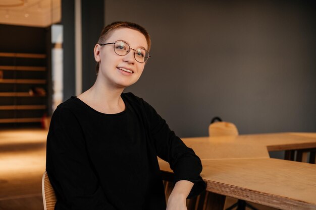 Lächelnde junge Frau mit kurzen Haaren, Brille und schwarzem Pullover, die im Büro am Tisch sitzt