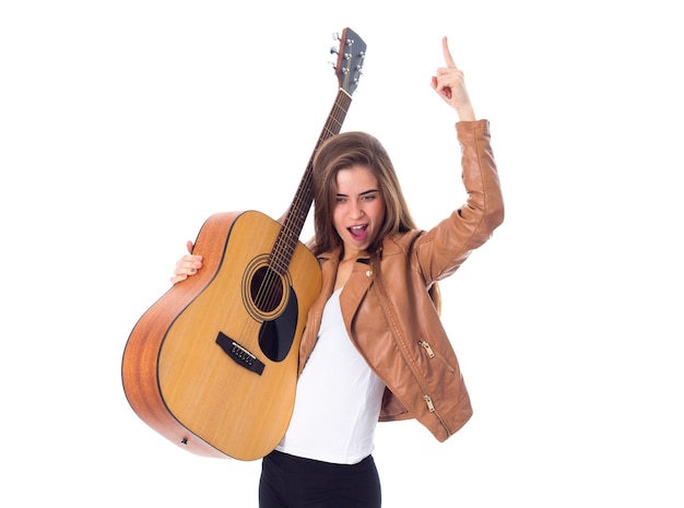 Lächelnde junge Frau mit langen Haaren in brauner Jacke und schwarzer Hose, die eine Gitarre hält und den Finger auf weißem Hintergrund im Studio zeigt