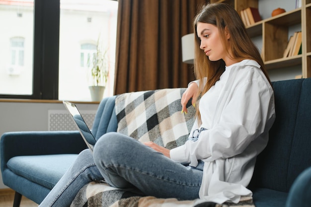 Lächelnde junge Frau mit Laptop, die zu Hause auf der Couch sitzt. Schönes Mädchen, das online in sozialen Netzwerken einkauft oder chattet und Spaß daran hat, freiberufliche Filme anzusehen, die an Computerprojekten arbeiten