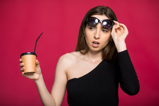 Lächelnde junge Frau mit Sonnenbrille, die isoliert vor rotem Hintergrund steht und eine Kaffeetasse zum Mitnehmen hält