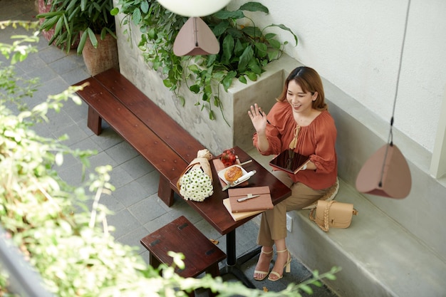 Lächelnde junge Frau mit Tablet-Computer, die am Café-Tisch sitzt und Freund winkt, der vorbeigeht?