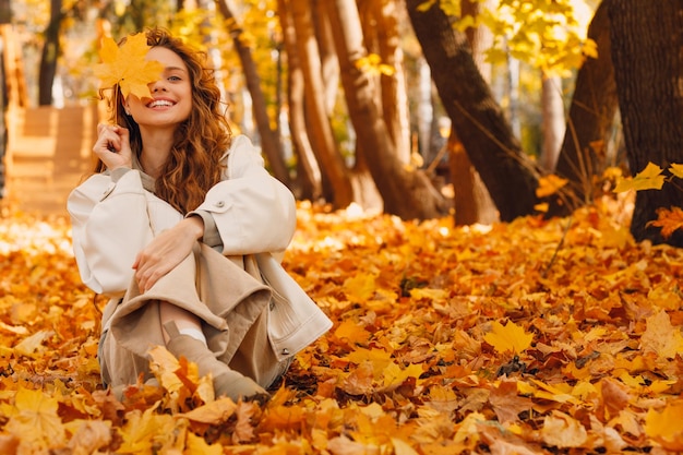 Lächelnde junge Frau sitzt im Herbstlaub auf dem Boden des Waldes mit den gelben Blättern