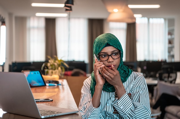 Lächelnde junge Frau spricht über Smartphone-Arbeit auf Laptop-Look im Ferndenken. Glücklicher afroamerikanischer Kundenkunde, der moderne Gadgets, Mobiltelefone und Computer im Heimbüro verwendet. Hochwertige Pho