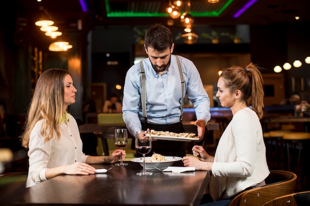 Lächelnde junge Freundinnen in einem Restaurant mit Kellner, der Abendessen dient