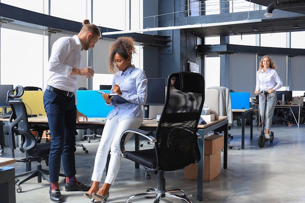 Lächelnde junge Geschäftsfrau, die während ihrer Arbeitspause mit einem Roller durch ein großes, modernes Büro fährt