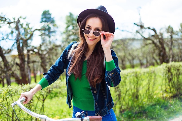 Lächelnde junge Hipster-Frau auf einem Fahrrad, die mit Sonnenbrille und Hut durch die Stadt fährt und Spaß hat