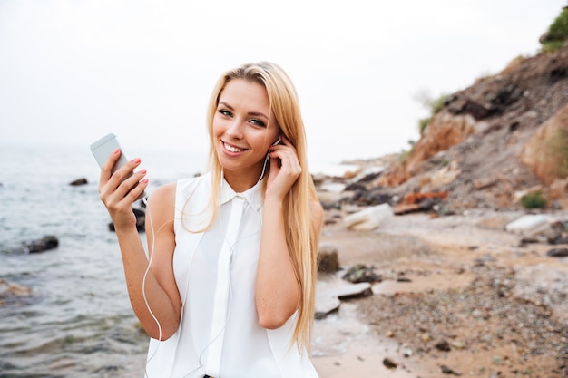 Lächelnde junge hübsche Frau, die Musik mit Smartphone hört, während sie am felsigen Strand steht