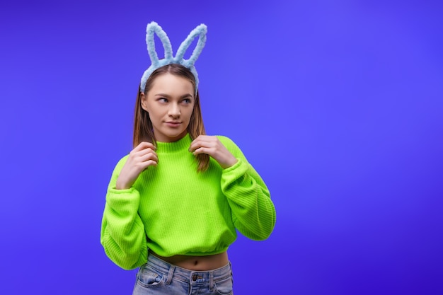 Lächelnde junge kaukasische Frau, Blondine mit blauen Hasenohren und einem grünen Pullover, Blick auf Kamera lokalisiert über blaue Wand