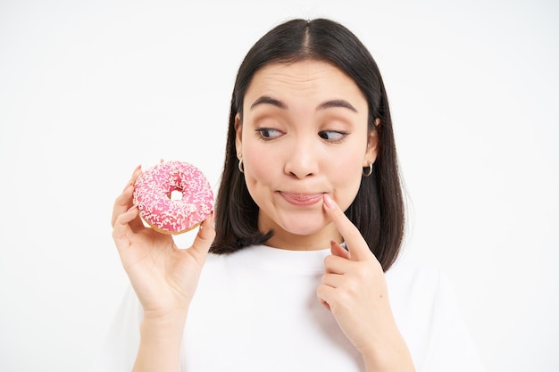 Lächelnde junge koreanische Frau denkt nach und blickt auf den rosa glasierten Donut-weißen Hintergrund