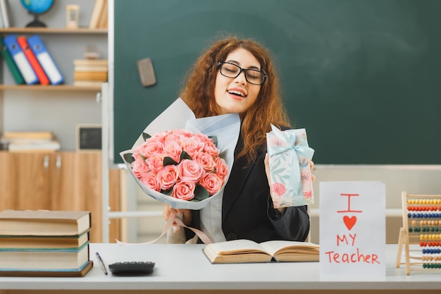 lächelnde junge lehrerin, die blumenstrauß mit geschenk hält, das am schreibtisch mit schulwerkzeugen im klassenzimmer sitzt