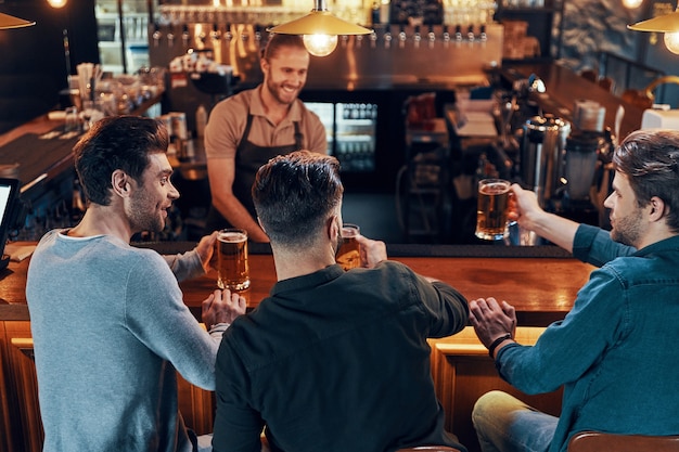 Lächelnde junge Männer in Freizeitkleidung trinken Bier, während sie zusammen in der Kneipe sitzen