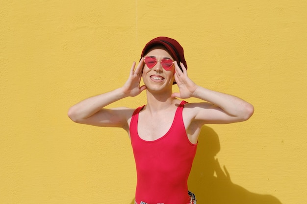 Lächelnde junge nicht-binäre Person mit Baskenmütze und Sonnenbrille, während sie im Freien gegen die gelbe Wand posiert.