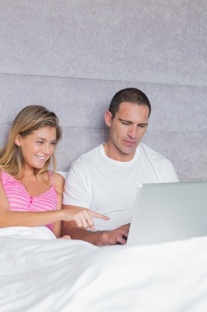 Lächelnde junge Paare, die zusammen ihren Laptop im Bett verwenden