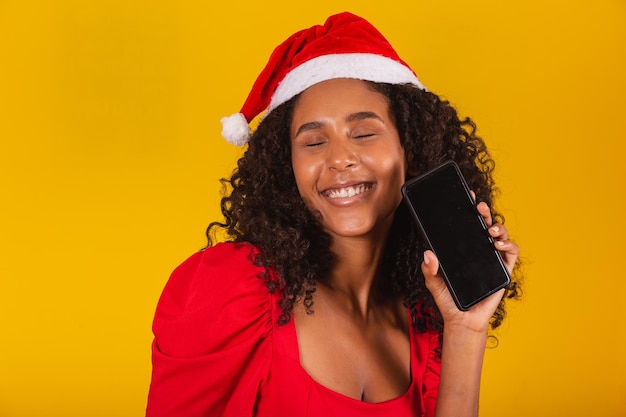 Lächelnde junge Sankt-Frau im Weihnachtshut, der Mobiltelefon mit leerem leerem Bildschirm lokalisiert auf gelbem Hintergrund hält.