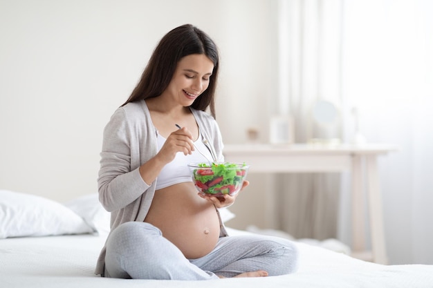 Lächelnde junge schwangere Frau, die zu Hause gesunden Salat isst