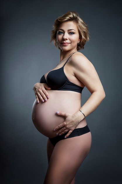 Lächelnde junge schwangere Frau in Unterwäsche berührt ihren Bauch mit ihren Händen. Liebe und Zärtlichkeit. Grauer Hintergrund. Vertikal.