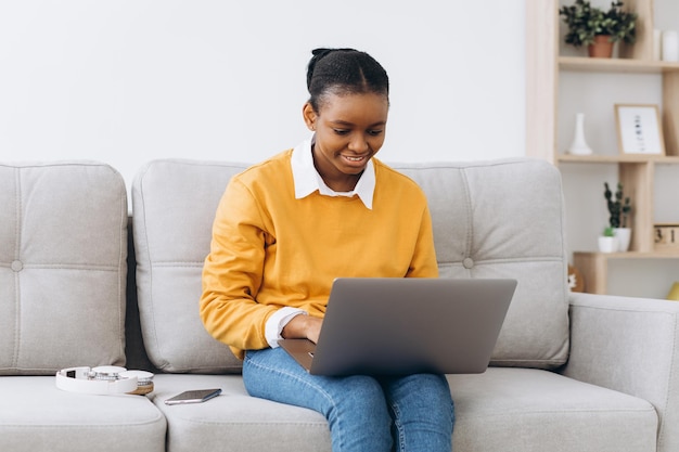 Lächelnde junge schwarze Frau im gelben Pullover, Freiberuflerin, die von zu Hause aus arbeitet, auf dem Sofa sitzt, Laptop benutzt, Platz kopiert und ernsthaft denkt.