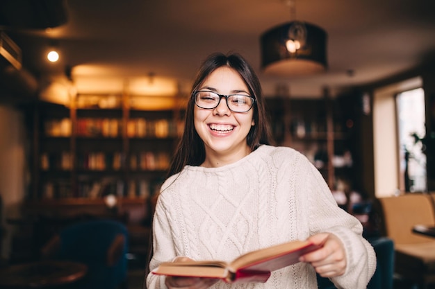 Lächelnde junge Studentin lacht und hält ein Buch in der Bibliothek