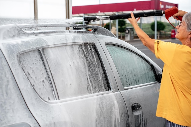 Lächelnde kaukasische Seniorin, die ihr Auto in einer Selbstbedienungs-Autowaschstation mit Hochdruckwasser wäscht Selbstbedienung in der Autowaschanlage