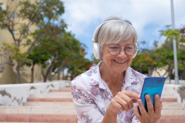 Lächelnde kaukasische Seniorin mit Kopfhörern und Brillen, die draußen auf einer Treppe in der Stadt mit dem Telefon sitzt