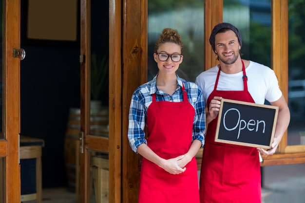 Lächelnde Kellnerin und Kellner stehen mit offenem Schild außerhalb des Kaffees