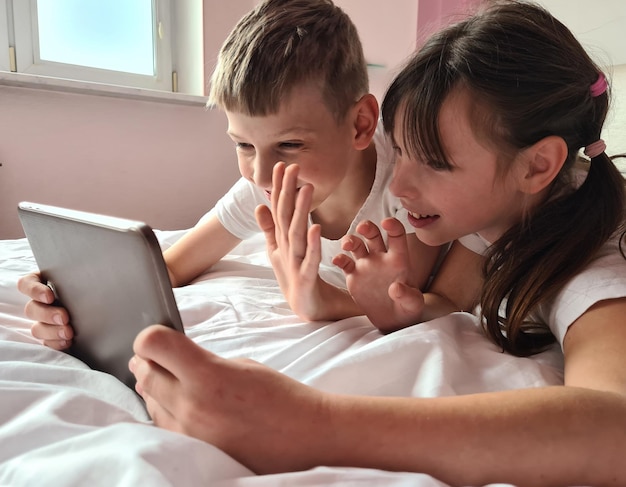 Lächelnde kleine Kinder, Junge und ein Mädchen, die im Bett liegen und auf den Bildschirm des Tablets schauen
