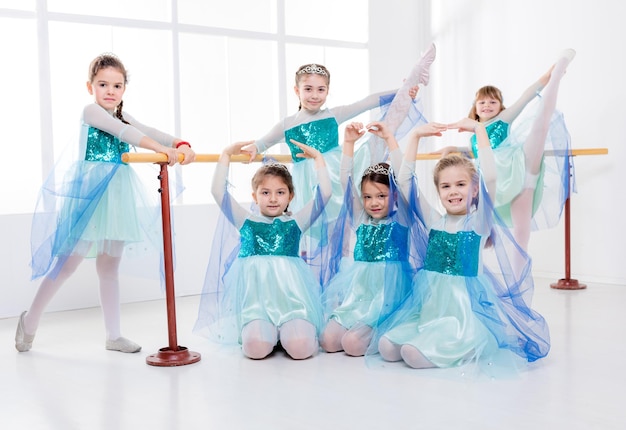 Lächelnde kleine Mädchen in Kleidern, die während des Ballettunterrichts Körperhaltungen üben. Blick in die Kamera.