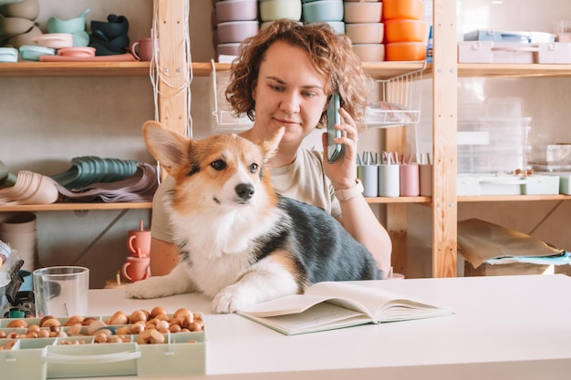 Lächelnde, konzentrierte Geschäftsfrau, die über ein Smartphone spricht, das einen süßen Hundecorgi in der Werkstatt hält Silikon-Babygeschirr-Shop